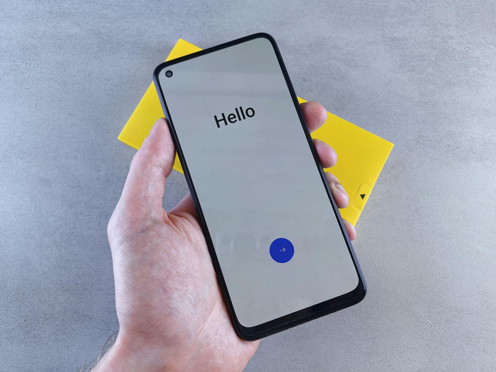 Dłoń trzymająca smartfon na szarej powierzchni obok żółtego pudełka