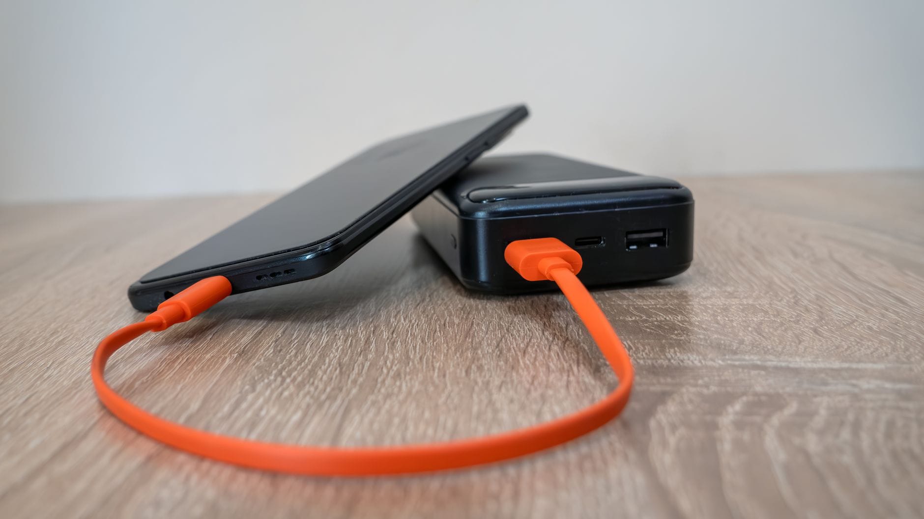 Smartfon ładowany z powerbanku pomarańczowym kablem na drewnie